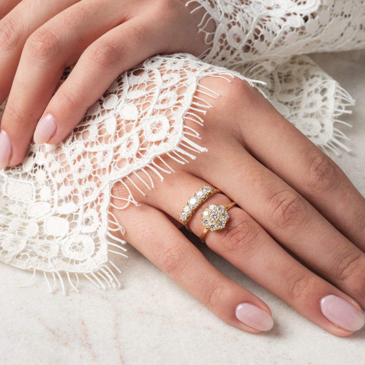 A close-up of a hand with light pink nails wearing two gold rings—an Anniversary Band with 0.78 ctw sparkling moissanite and a floral diamond cluster ring—resting on delicate white lace fabric.