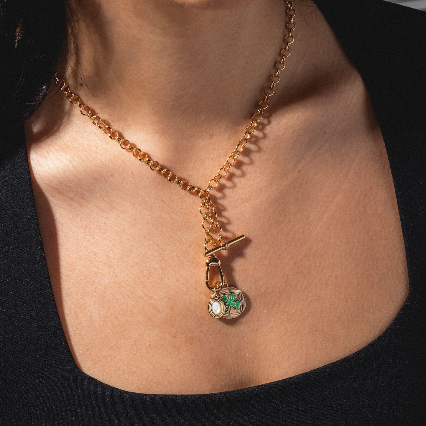 A close-up of a person wearing the Monroe Birthstone Charm—a gold chain necklace with a toggle clasp and a round pendant featuring a green and blue butterfly design—against a black top.