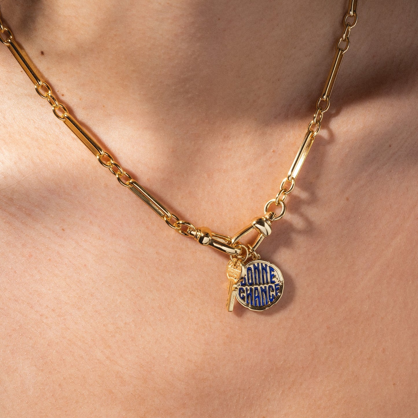 A close-up of the Liam Watch Chain Charm Holder Necklace, featuring a small gold key charm and a round pendant inscribed with BONNE CHANCE in blue, worn around a person’s neck.