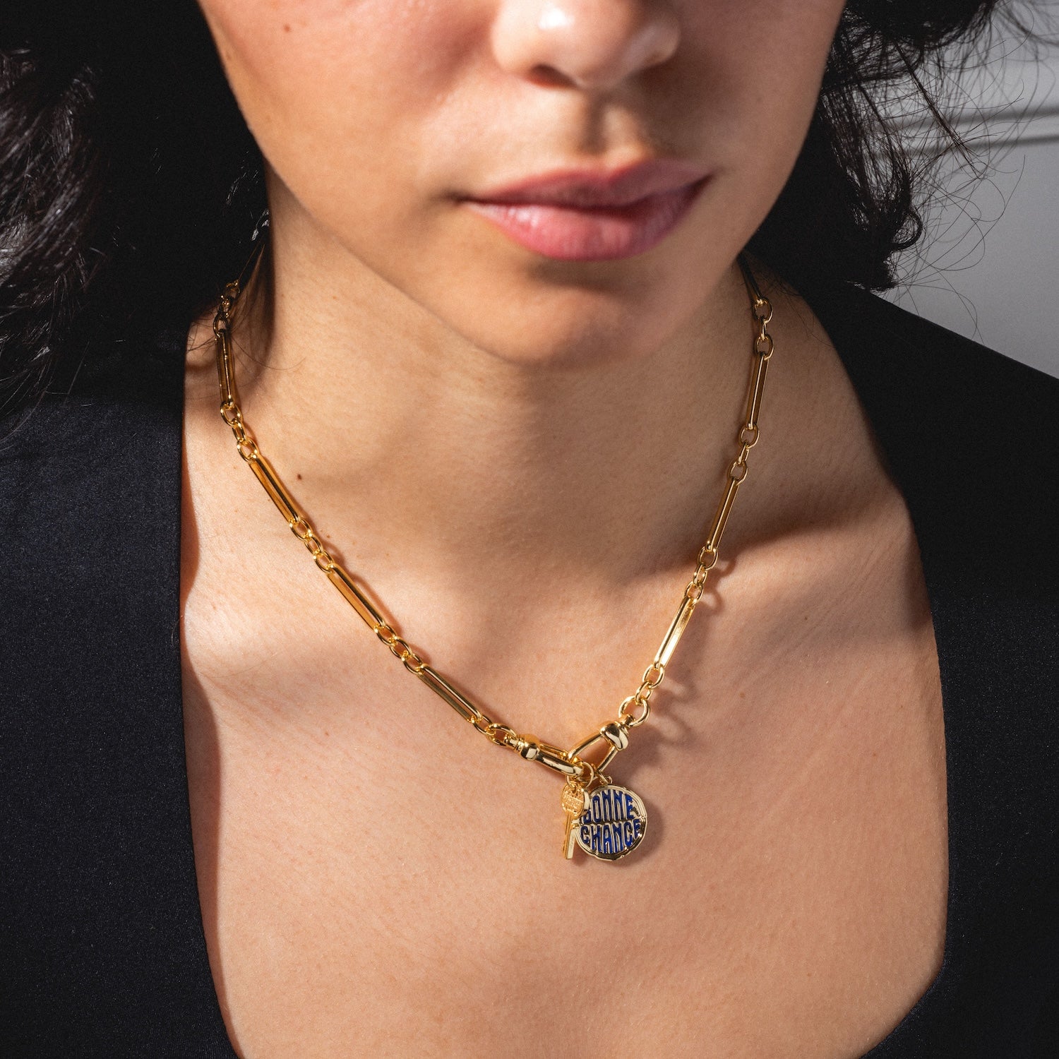 A woman dressed in a black top wears the Liam Watch Chain Charm Holder Necklace, featuring a round pendant inscribed with "GAME CHANGER." She is shown from the shoulders up.