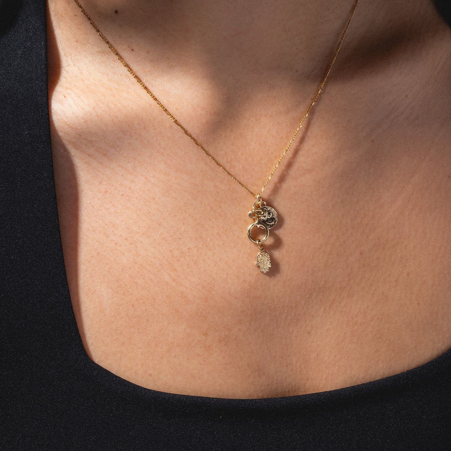 A close-up of a person wearing the Charms by Charlie and Marcelle gold necklace, featuring multiple delicate charms including a small round pendant, against bare skin and a black top.