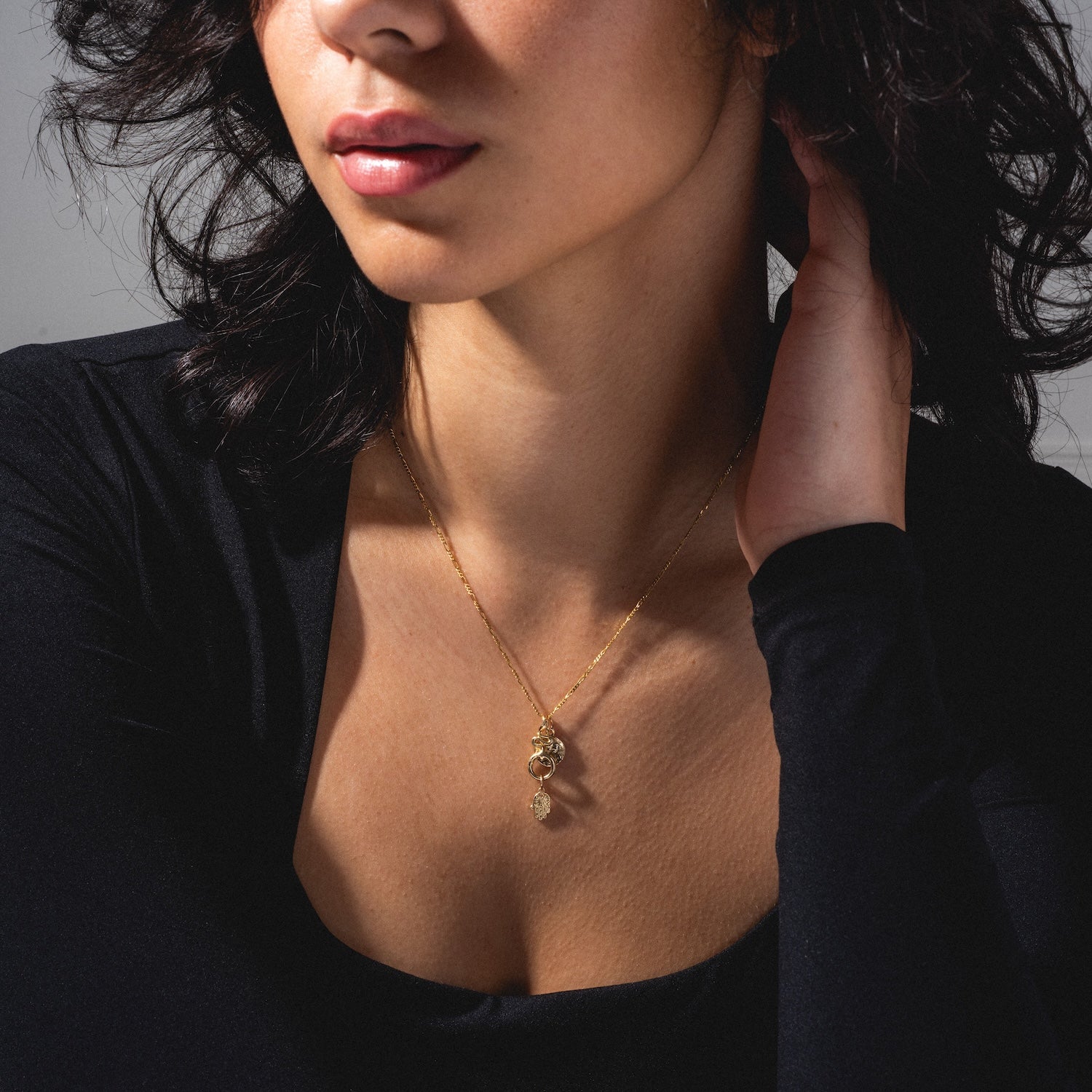 A woman with wavy dark hair in a black top touches her neck, showcasing two layered Charms by Charlie and Marcelle gold necklaces with small pendants against a neutral background.