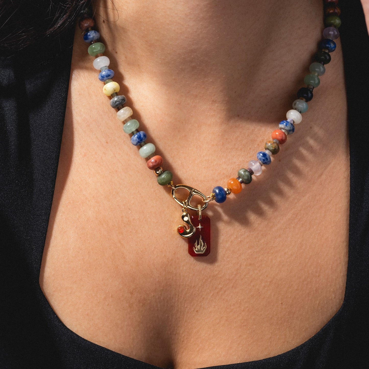 A close-up of a person's neck wearing Thea Bubble Letter Charm on a colorful beaded necklace with a gold clasp, red charm, and gold flame-shaped pendant, paired with a black top.