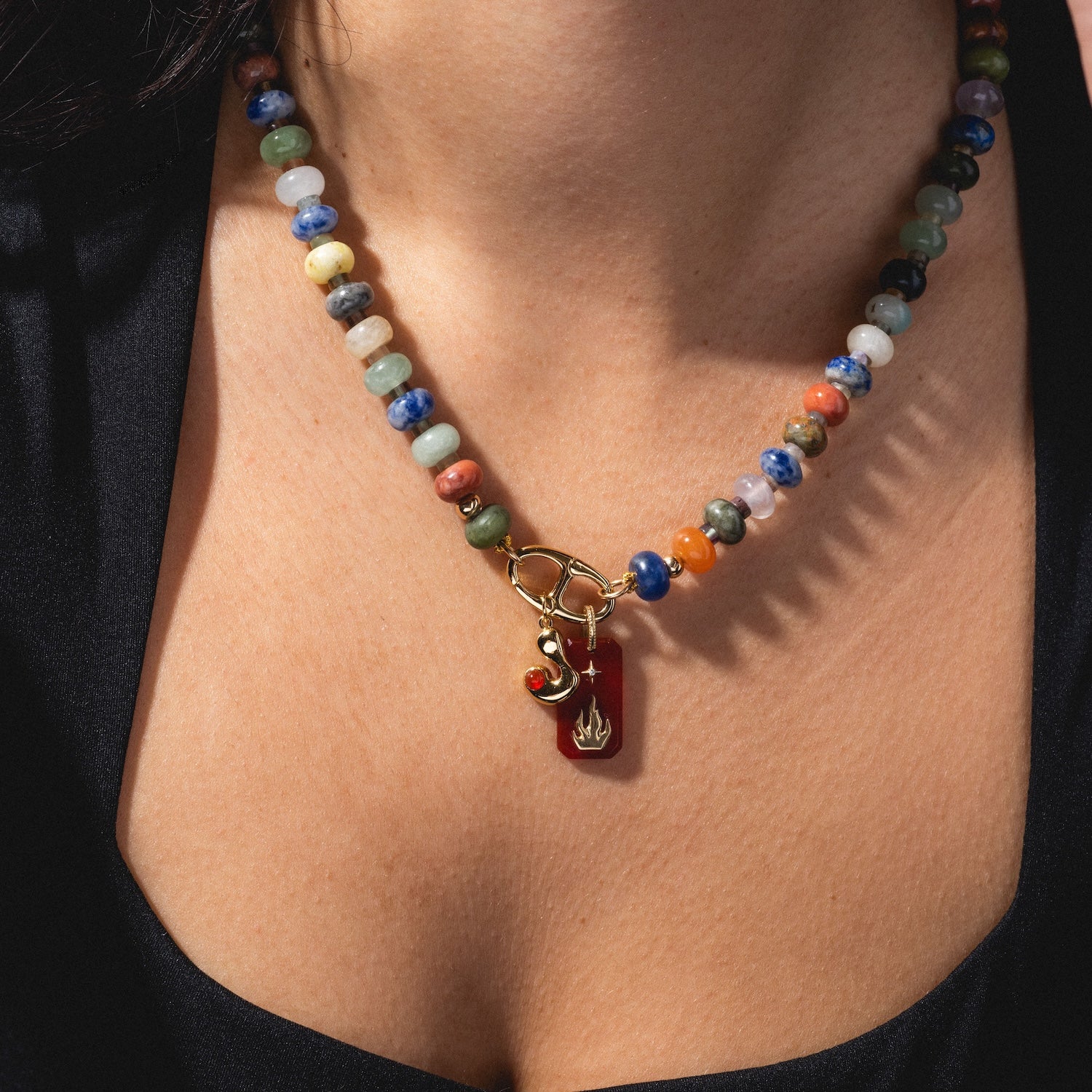 A close-up of a person's neck wearing Thea Bubble Letter Charm on a colorful beaded necklace with a gold clasp, red charm, and gold flame-shaped pendant, paired with a black top.