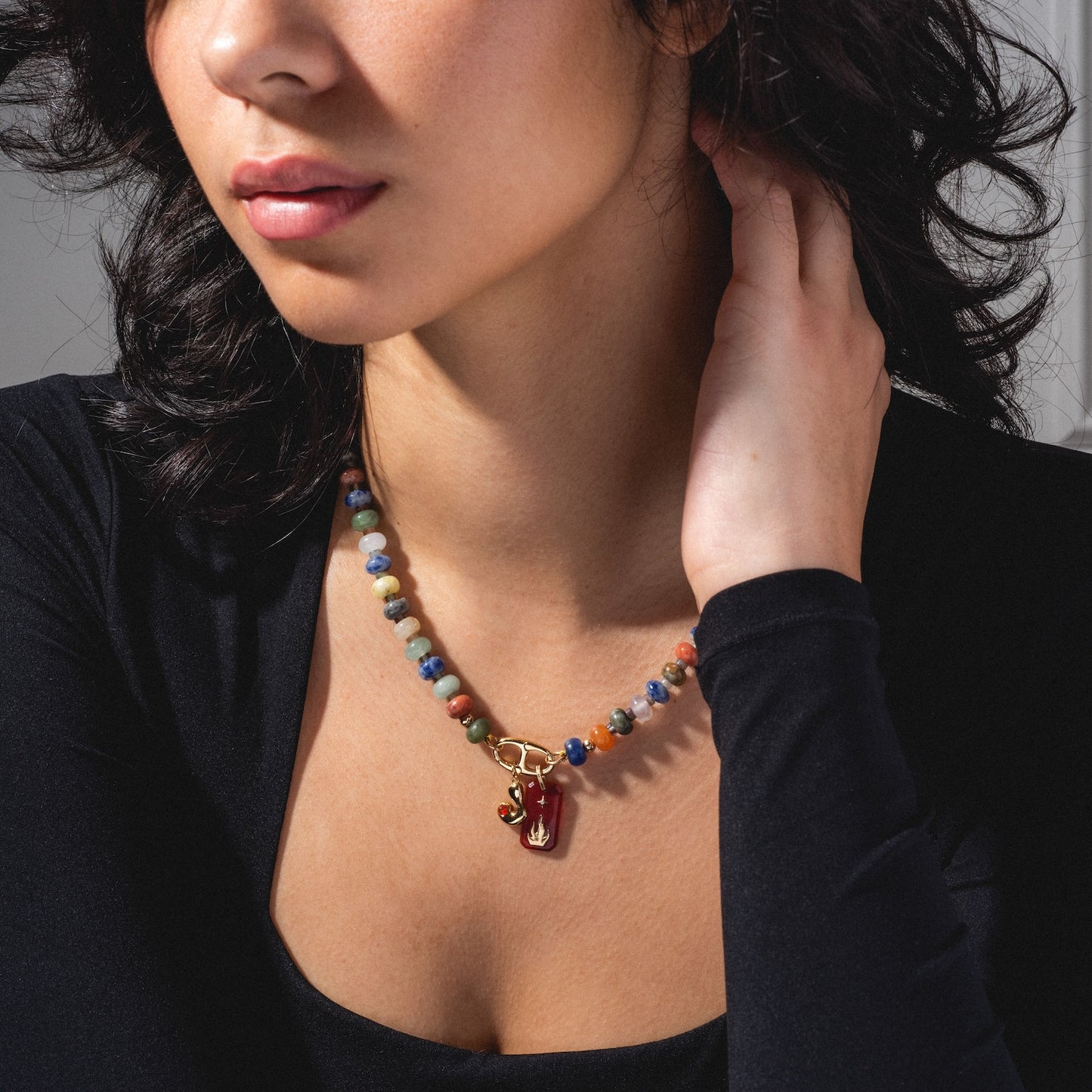 A woman with wavy dark hair wears a black top and the Macey Customizable Beaded Charm Holder Necklace, featuring a gold clasp and red pendant, as she rests her hand by her neck.