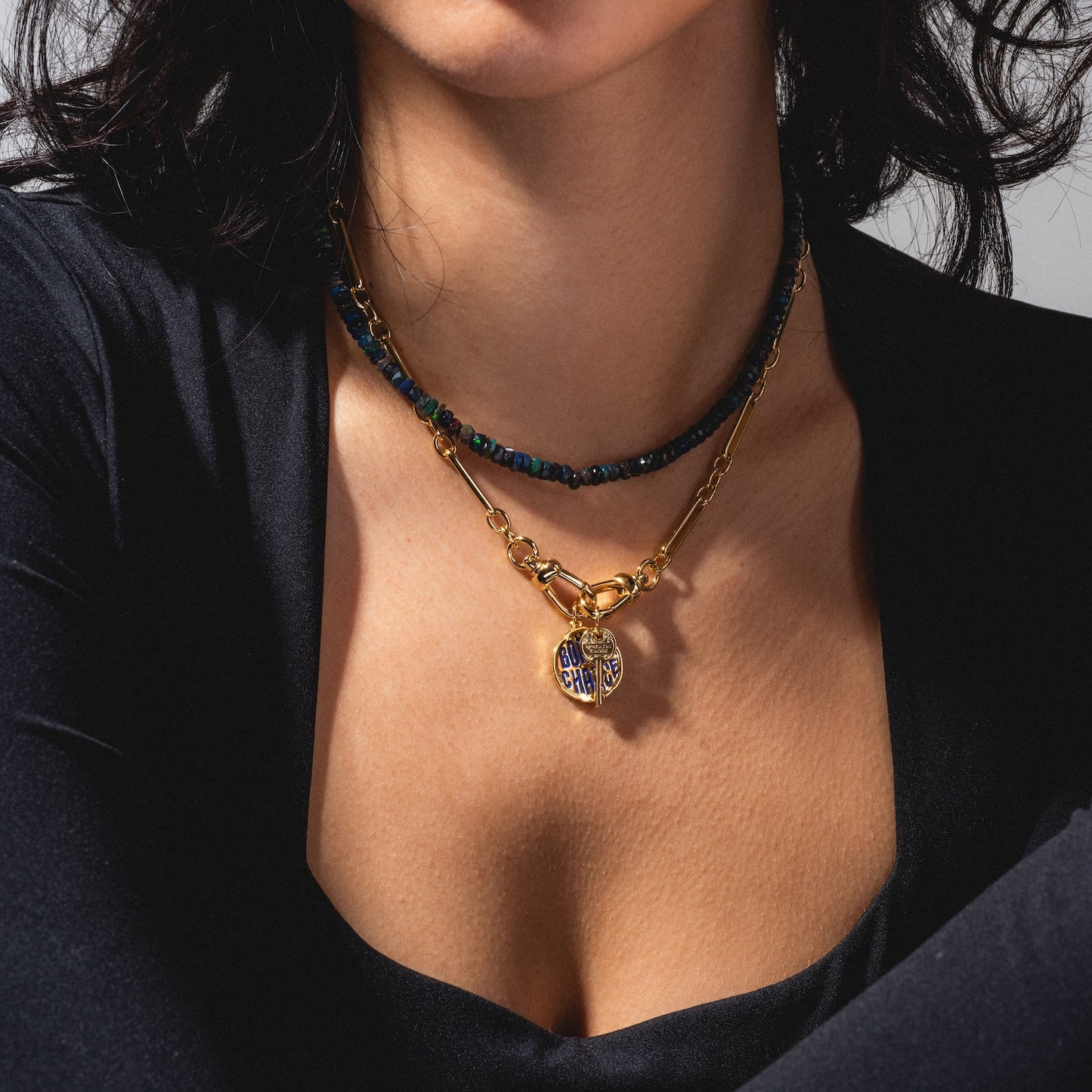 A woman wearing a black, low-cut top showcases two layered necklaces: one with dark beads, and the other—the Liam Watch Chain Charm Holder Necklace—featuring a round gold and blue pendant. The photo highlights her neck and collarbone.