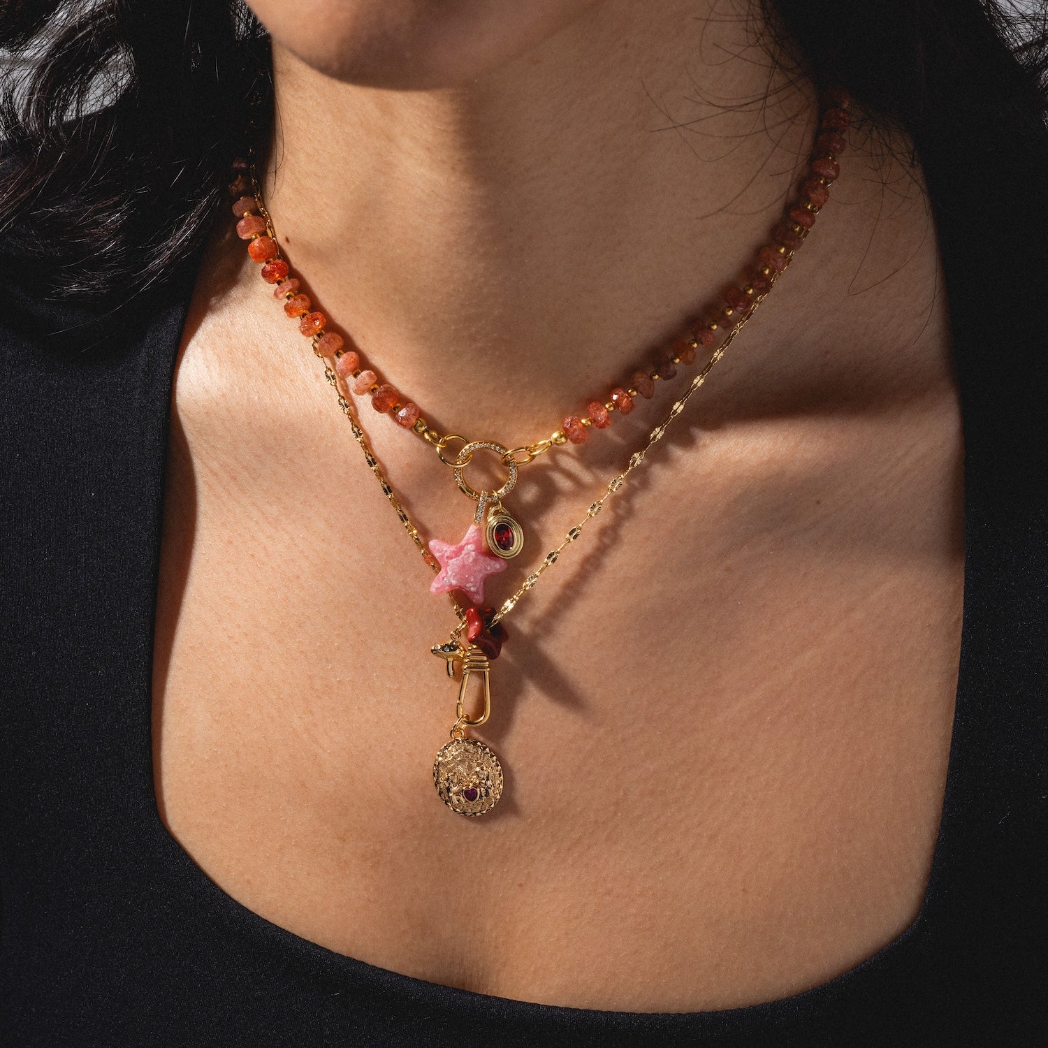 A woman wearing Gemstone Spacer Charms by Mod and Jo: three layered necklaces with orange beads, a pink star charm with gold pendant, and a gold medallion, styled over a black top.