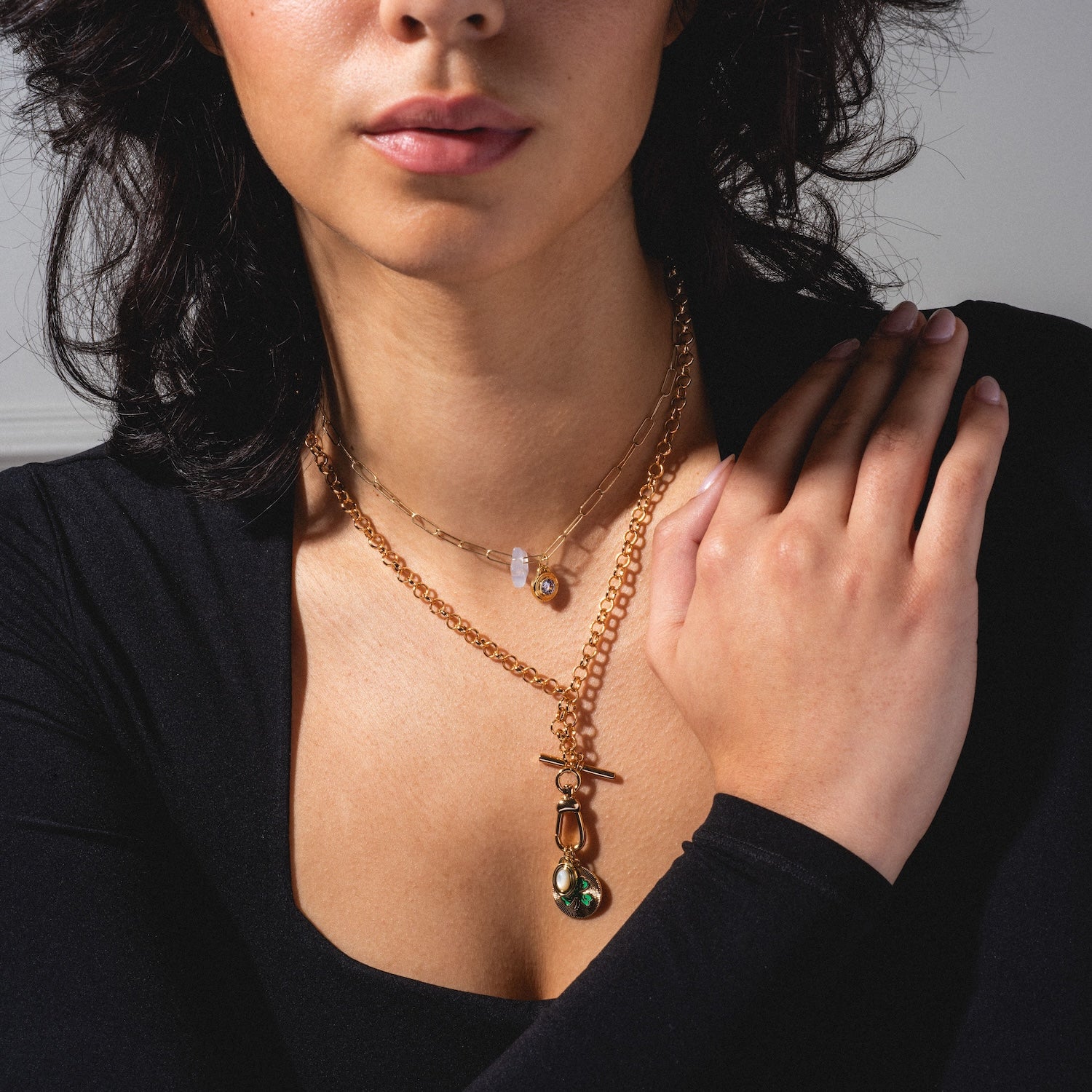 A woman in a black top displays Mod and Jo's Gemstone Spacer Charms layered as gold necklaces with pendants, including one with a green stone, her hand gently resting on her chest.