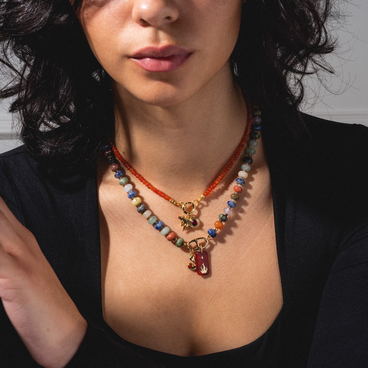 A woman with wavy dark hair wears a black top and three layered beaded necklaces featuring the Monroe Birthstone Charm with a red pendant, shown from shoulders to lips against a neutral background.