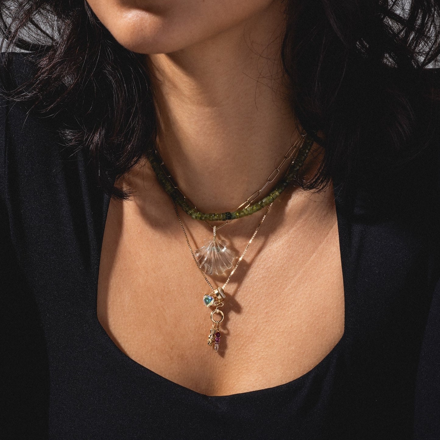 A woman wears Charms by Charlie and Marcelle: a green beaded choker, a gold chain with a clear shell pendant, and a gold chain with colorful charms. She pairs the layered necklaces with a black top; her dark hair is partially visible.