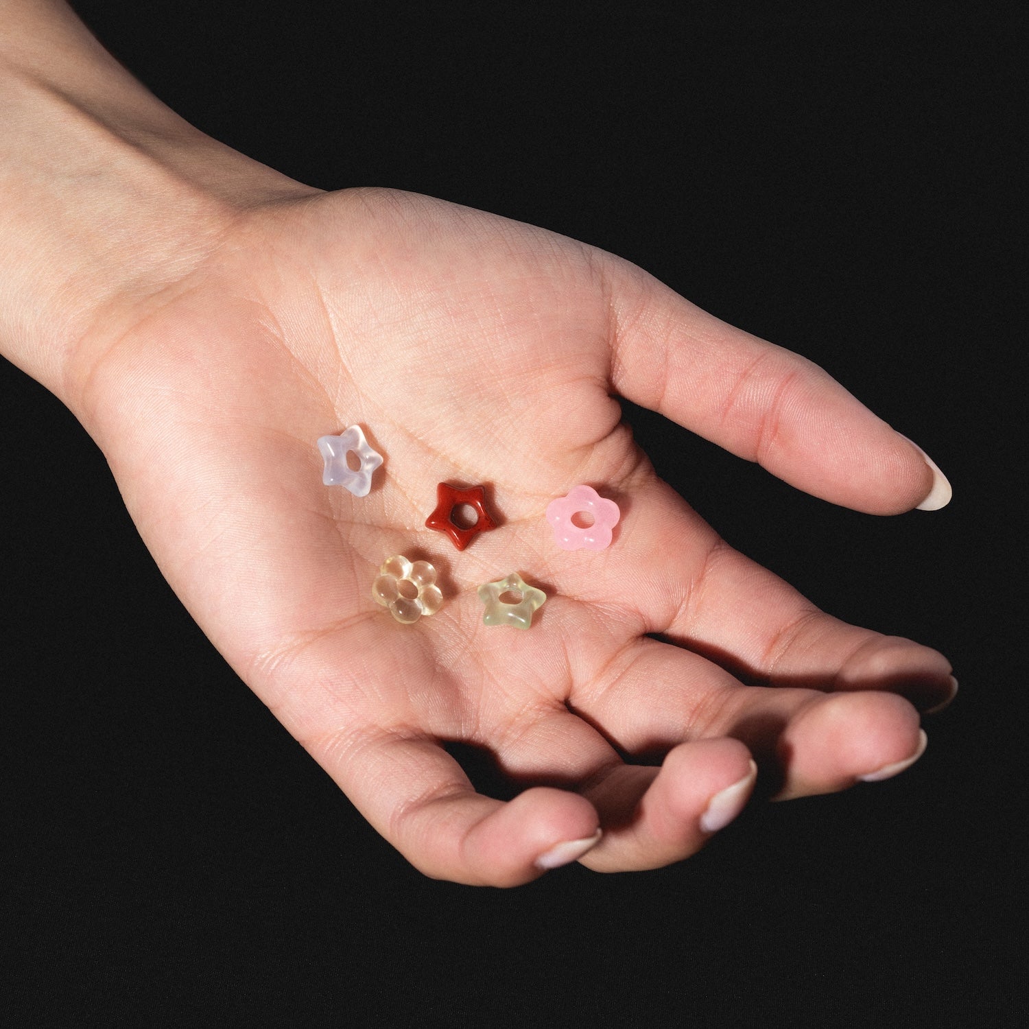 A hand with pale skin holds five Gemstone Spacer Charms by Mod and Jo, flower-shaped beads in white, red, pink, beige, and light yellow, against a black background.