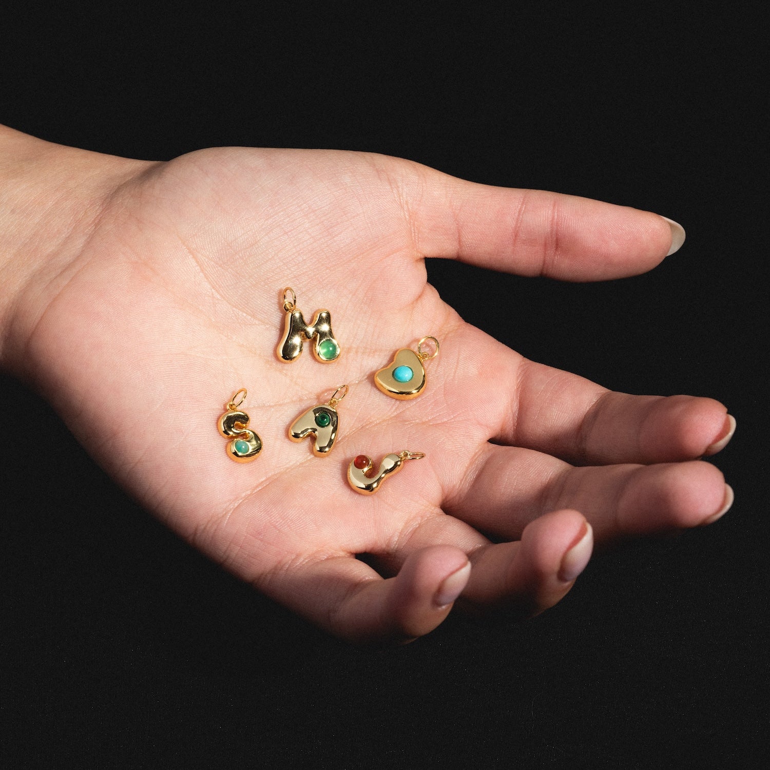 A hand holds five Thea Bubble Letter Charms with colorful stones, each gold pendant shaped as a different letter or symbol, set against a black background.