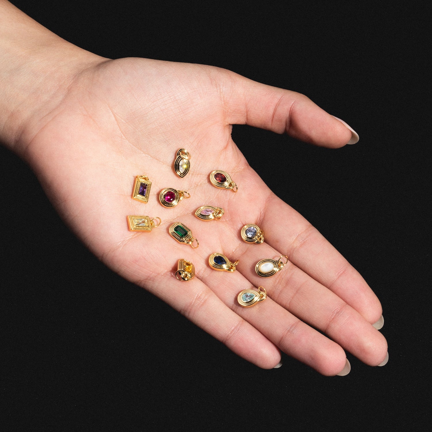 An open hand with a light skin tone holds twelve Monroe Birthstone Charms, each adorned with a different colored gemstone, against a black background.