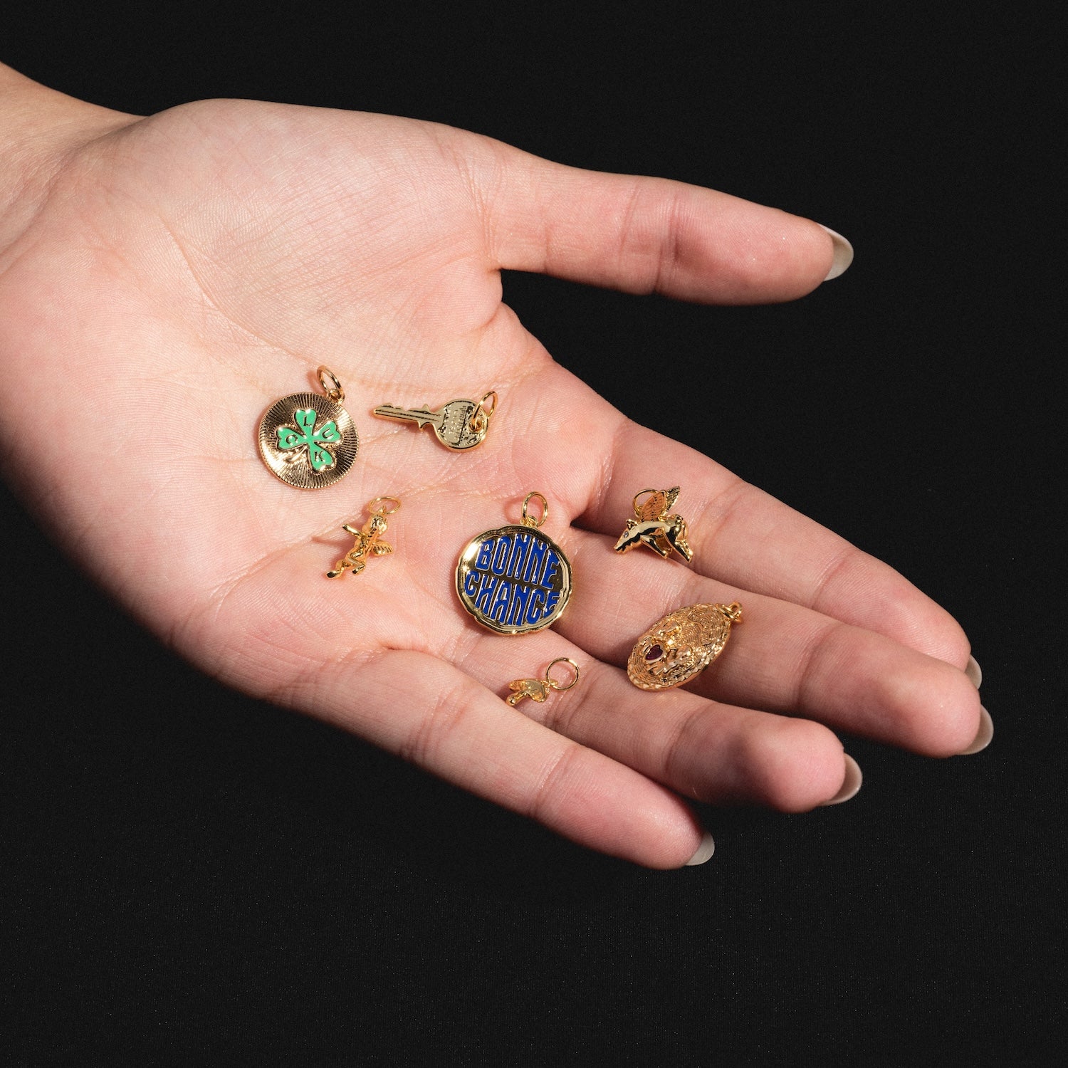 A manicured hand displays several Charms by Mod and Jo—such as a green four-leaf clover, blue BONNE CHANCE disk, key, and bird—against a black background.