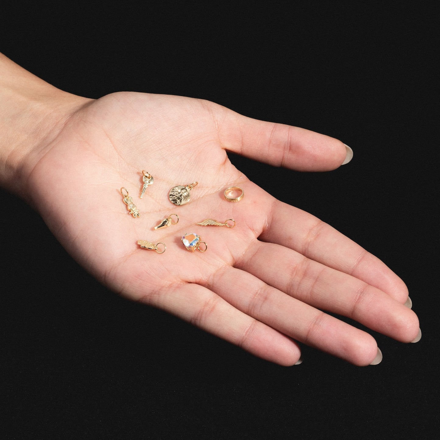 A hand with long nails holds several Charms by Charlie and Marcelle—small gold and crystal pieces—on its palm, set against a black background.