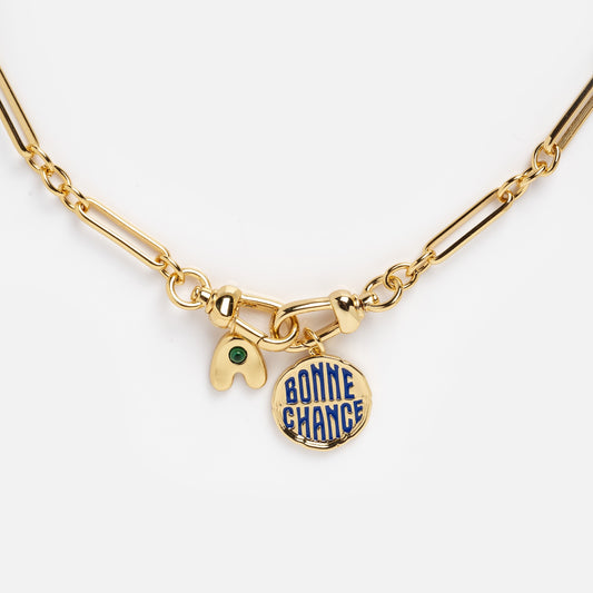 The Liam Watch Chain Charm Holder Necklace features a gold chain with two charms: a gold wishbone with a green gem and a round gold pendant inscribed with BONNE CHANCE in blue.
