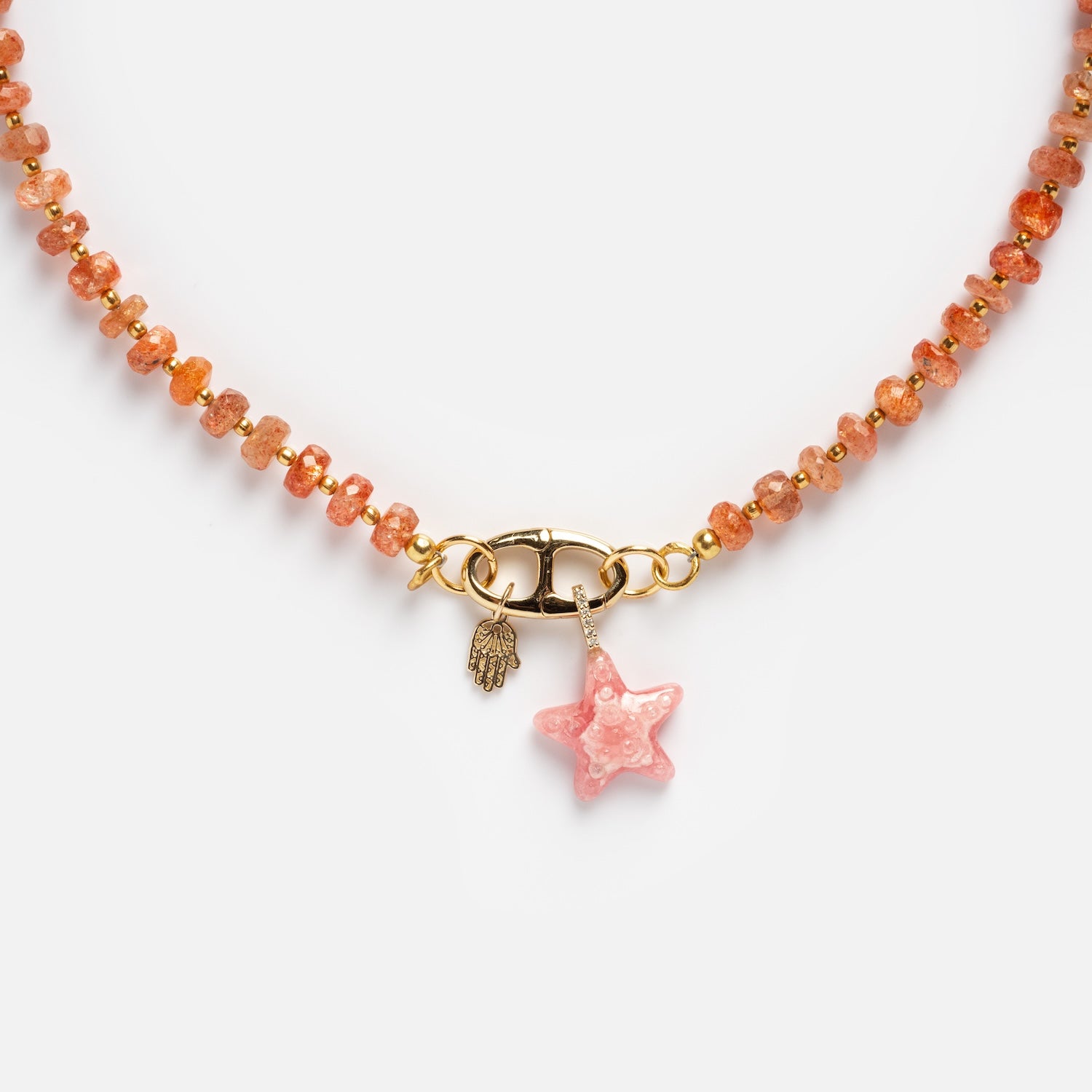 The Customizable Beaded Gemstone Charm Holder Necklace features translucent orange beads, a gold clasp, a small gold hamsa charm, and a pink star-shaped pendant at the center.