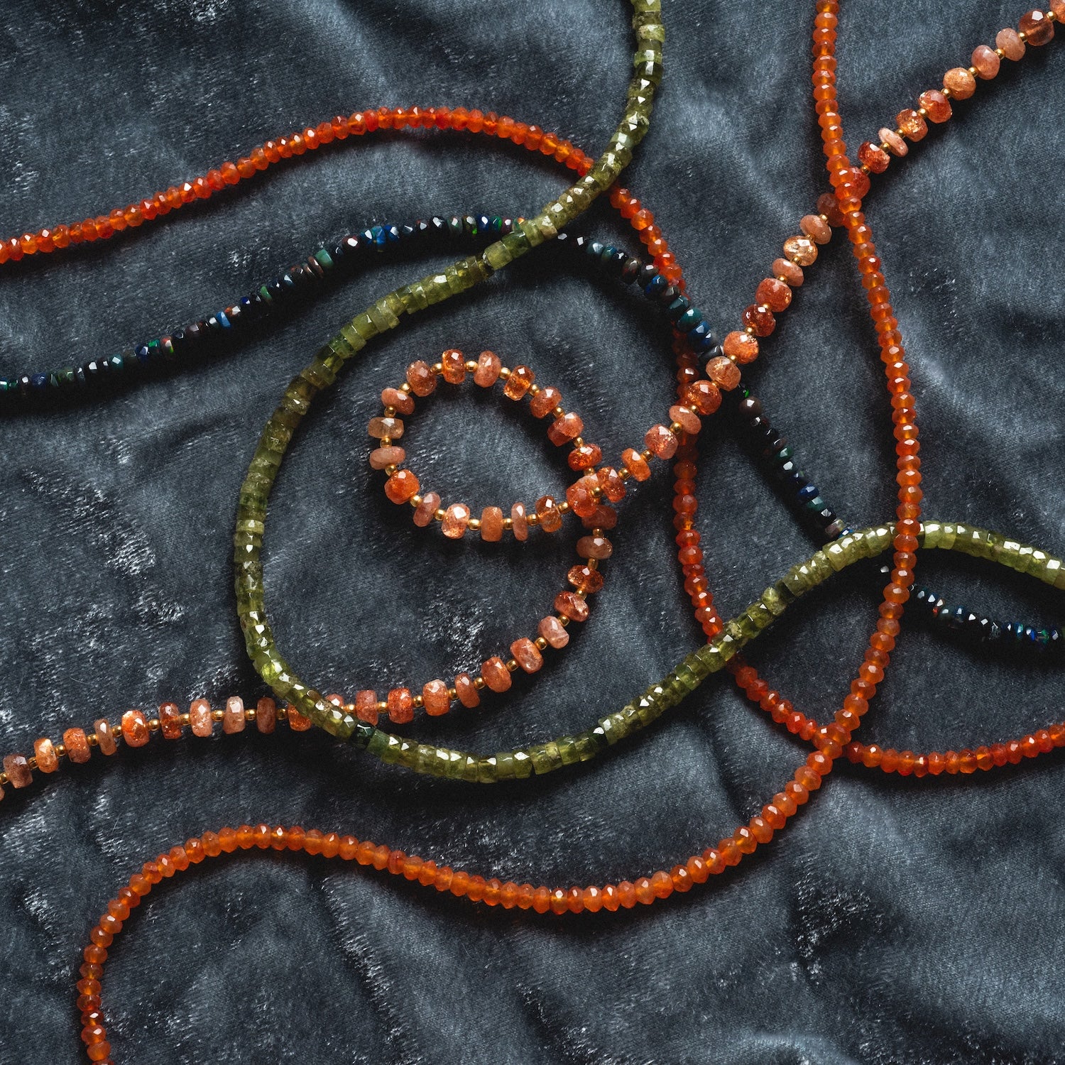 Several strands of orange, olive green, and black gemstone beads are loosely intertwined on a dark textured fabric, highlighting the Customizable Beaded Gemstone Charm Holder Necklace.