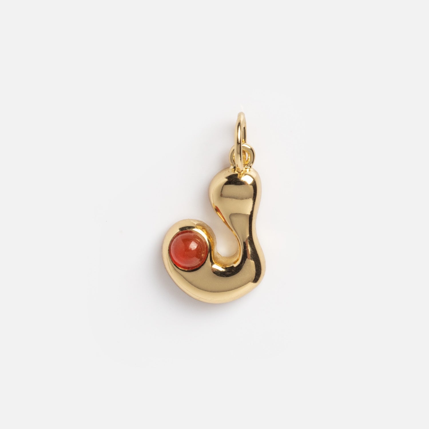 The Thea Bubble Letter Charm is a shiny, gold, abstract-shaped pendant featuring a small round red gemstone set in one curve, displayed against a plain white background.