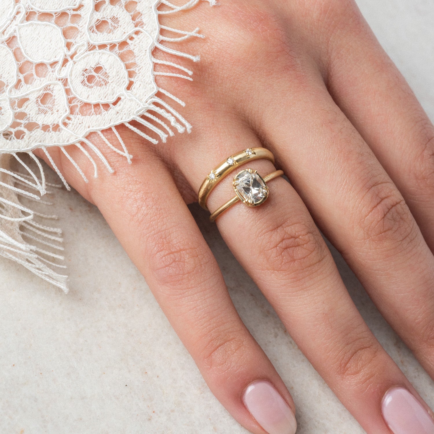 A hand with neatly manicured nails wears the Kismet Ring—a gold band with a large oval gemstone and natural diamonds, 1.3 mm wide—resting on a light surface. The sleeve is white lace with fringe.