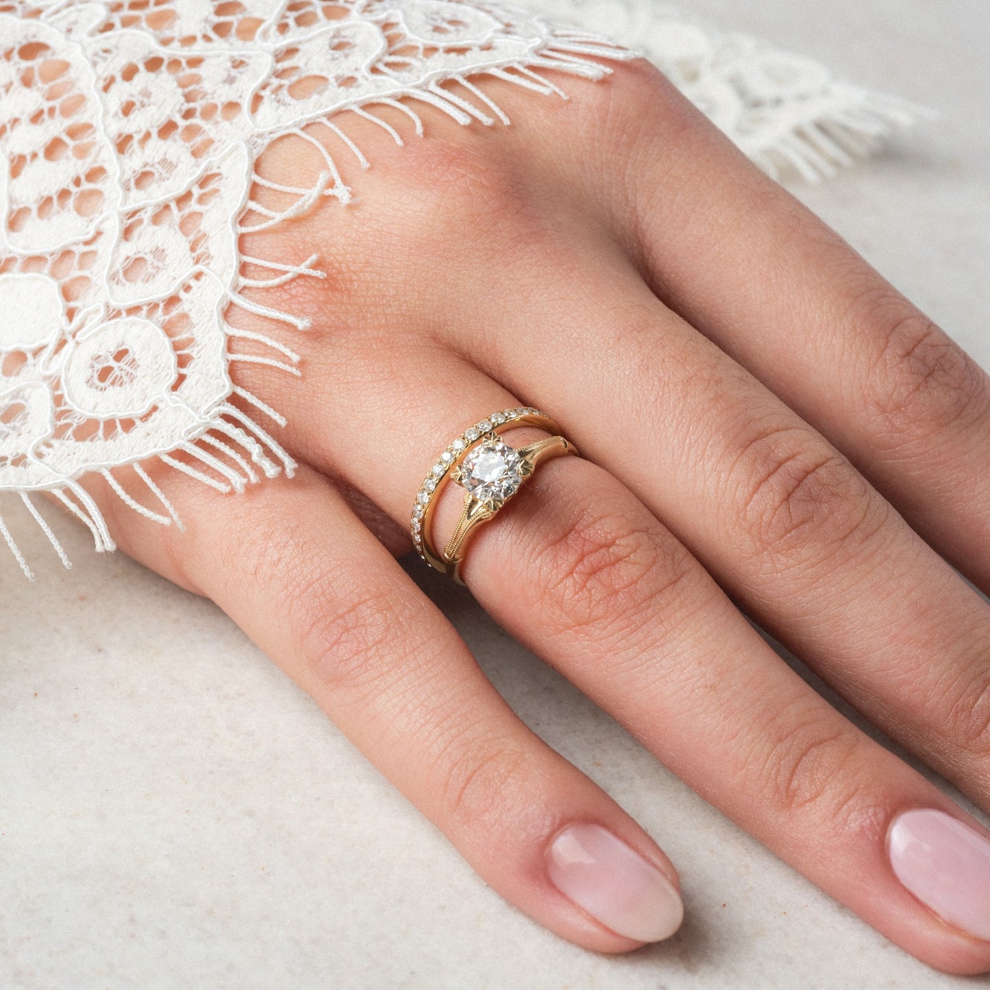 A hand with a light pink manicure wears two stacked gold rings: the Elara Ring, featuring a large round diamond, and a 1.4mm diamond band with 1.05 ctw of sparkling stones. The hand rests on a light surface, partially covered by white lace fabric.