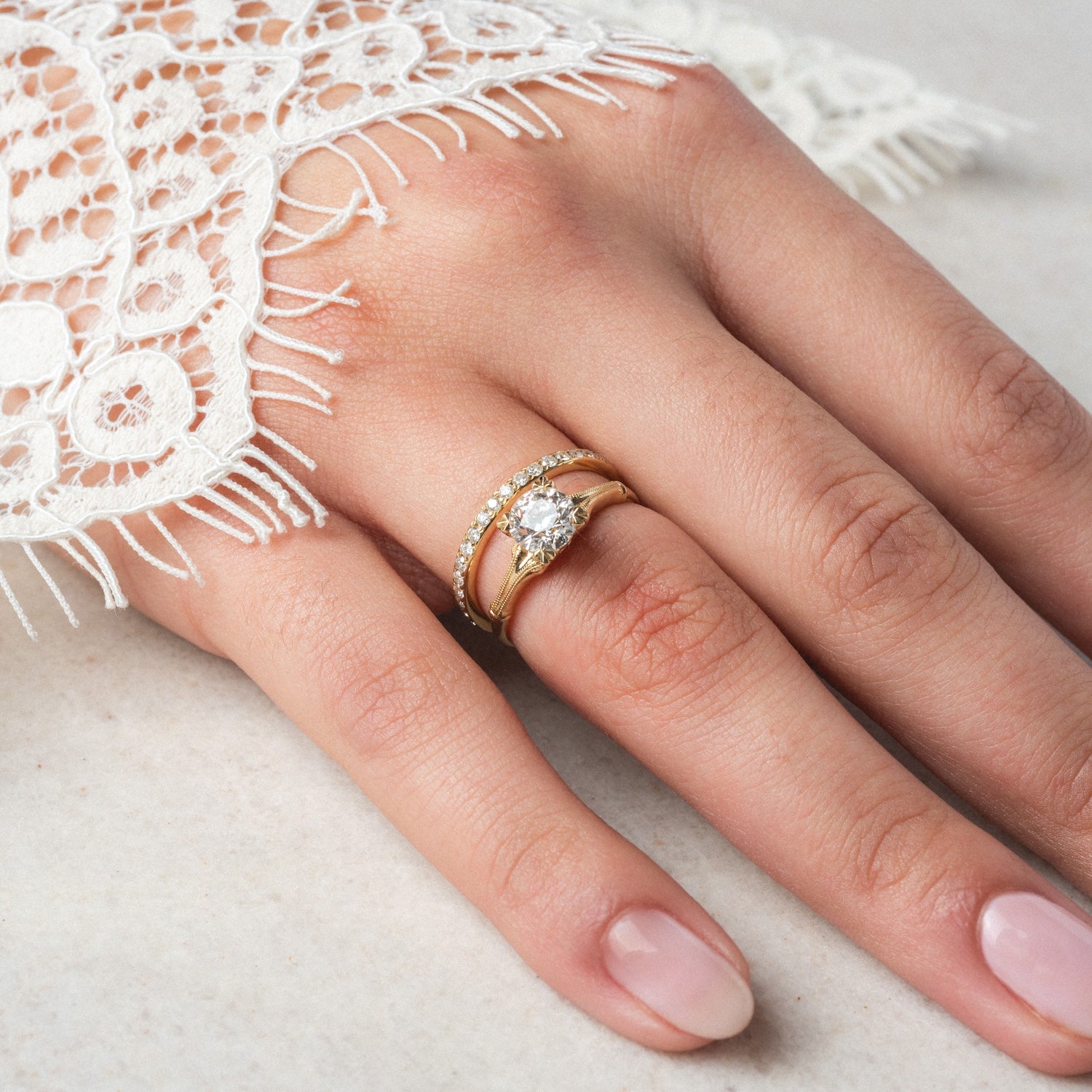 A hand with a light pink manicure wears two stacked gold rings: the Elara Ring, featuring a large round diamond, and a 1.4mm diamond band with 1.05 ctw of sparkling stones. The hand rests on a light surface, partially covered by white lace fabric.