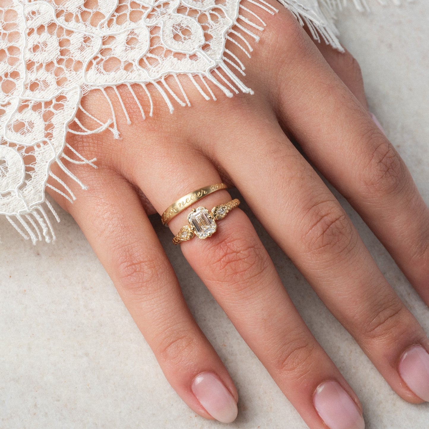 A hand with manicured nails wears the True Love Never Dies Band, a slim 1.2 mm gold ring with engraved text, alongside another gold ring featuring a large rectangular gemstone and smaller stones. Lace fabric partly covers the hand.