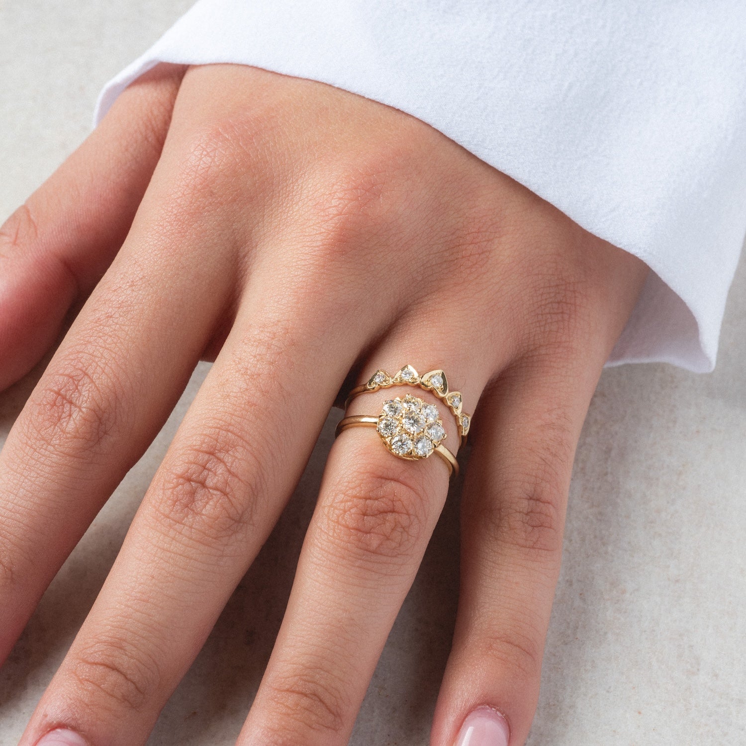 A hand wears three stacked Amas d'Etoiles Rings: one features a floral diamond cluster, the others have .86 ctw of small diamonds in geometric settings. The hand rests on a light surface, with a white sleeve visible.