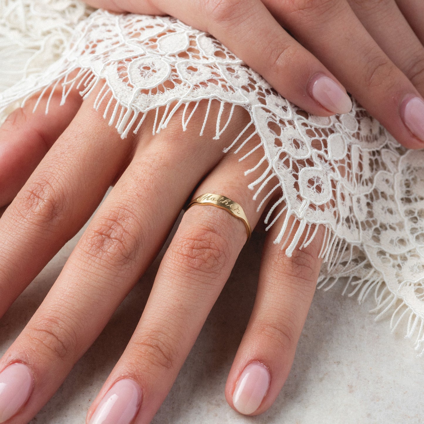 A close-up of a manicured hand wearing the 14k Ripples Customizable ID Band, engraved with text and partially covered by white lace fabric.