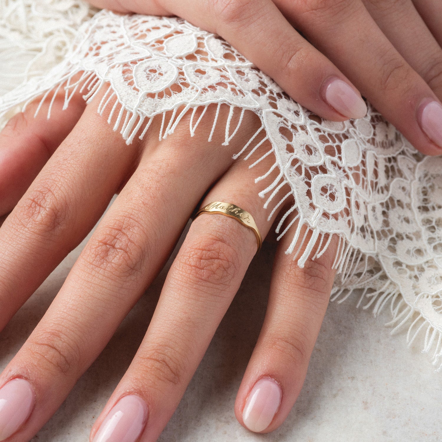 A close-up of a manicured hand wearing the 14k Ripples Customizable ID Band, engraved with text and partially covered by white lace fabric.