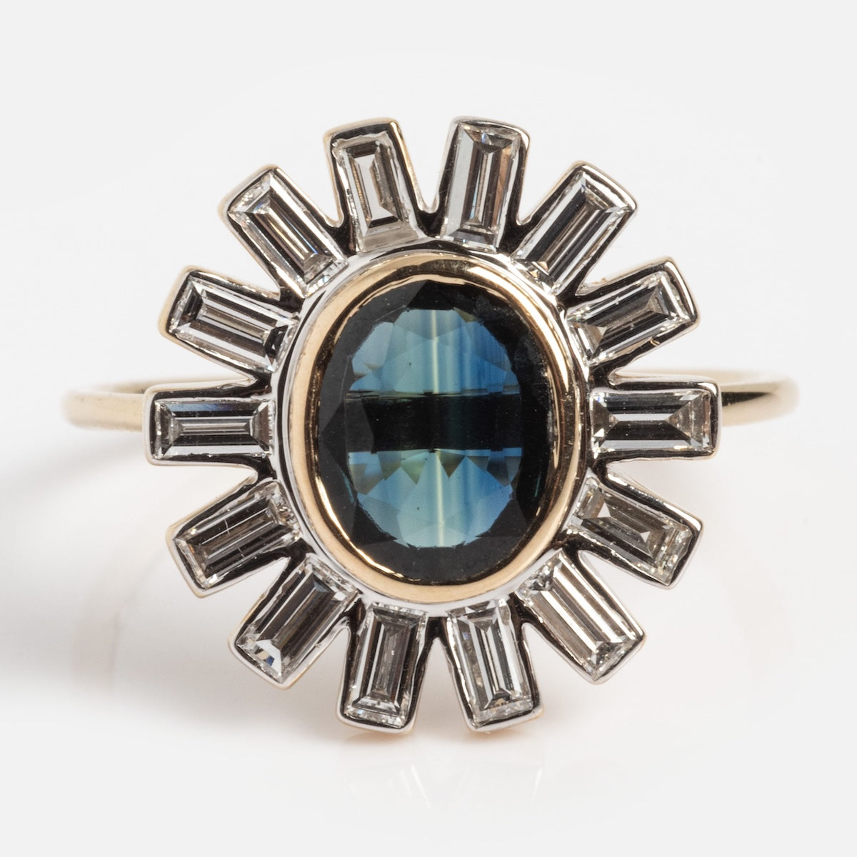 The Solid Gold Blue Sapphire Halo Ring (Sample Size 7) features a central oval blue sapphire, encircled by a sunburst of rectangular lab diamonds in silver-tone metal, shown on a white background.