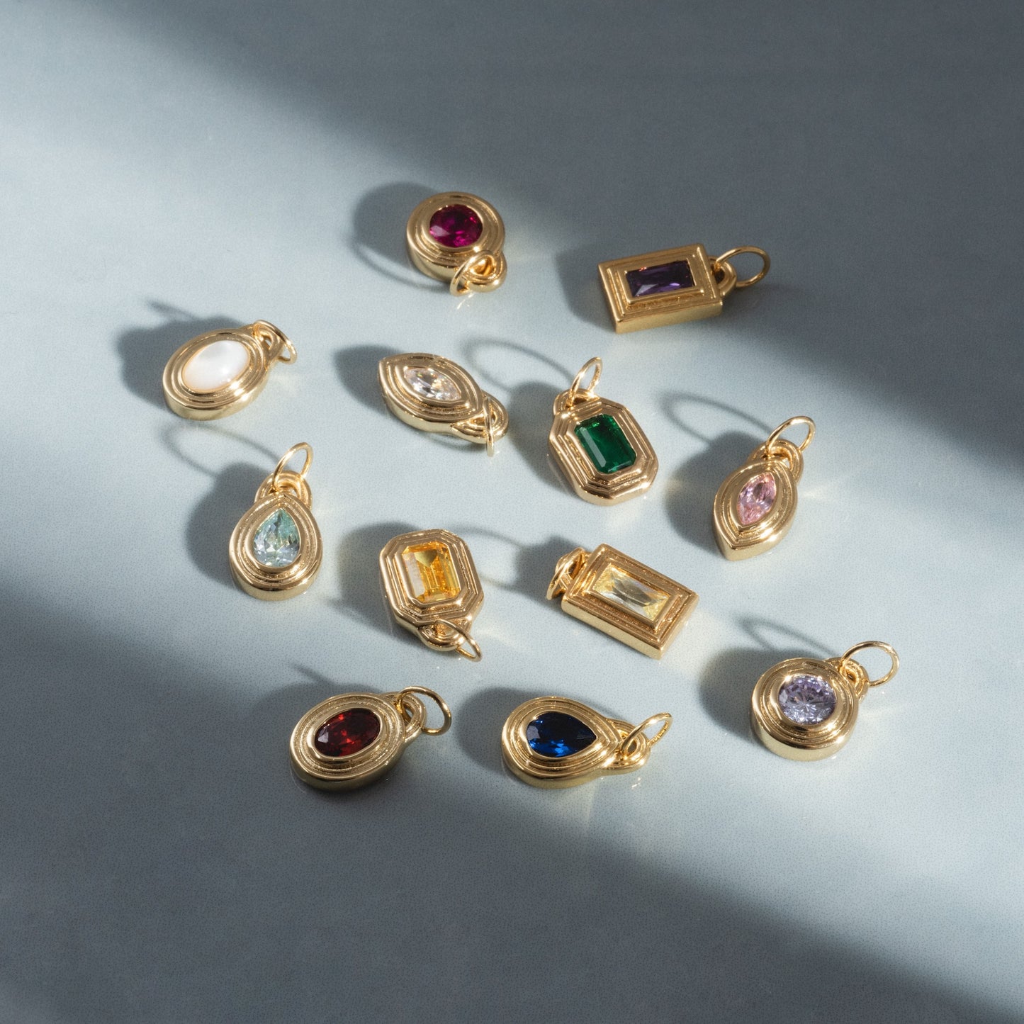 The Monroe Birthstone Charm, a gold pendant available in oval, round, and rectangular shapes with colorful gemstones, is displayed on a gray surface with soft sunlight and shadows.