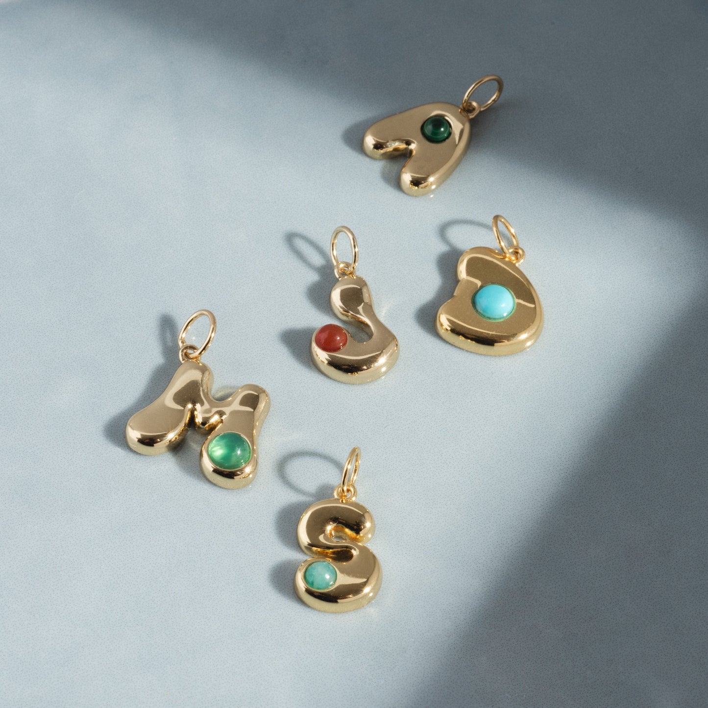 Five Thea Bubble Letter Charms in gold, featuring green, red, and turquoise stones in the letters M, S, A, L, and G, are arranged on a light blue background with soft shadows.