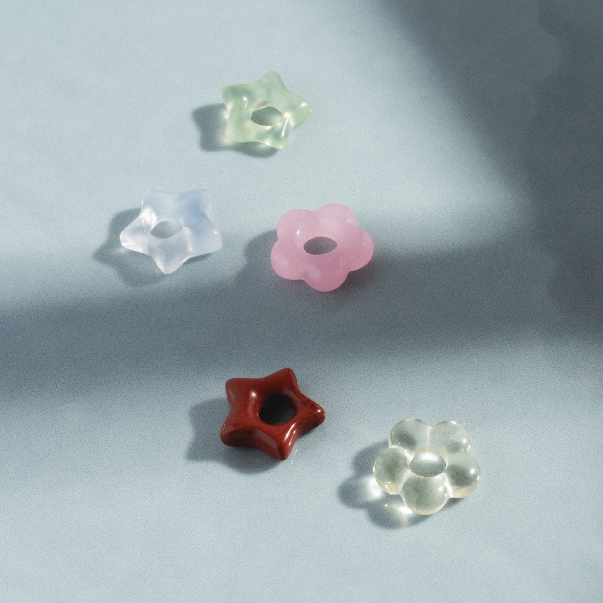 Five translucent, flower-shaped Gemstone Spacer Charms by Mod and Jo and one red star-shaped charm are arranged on a pale blue surface with soft shadows.