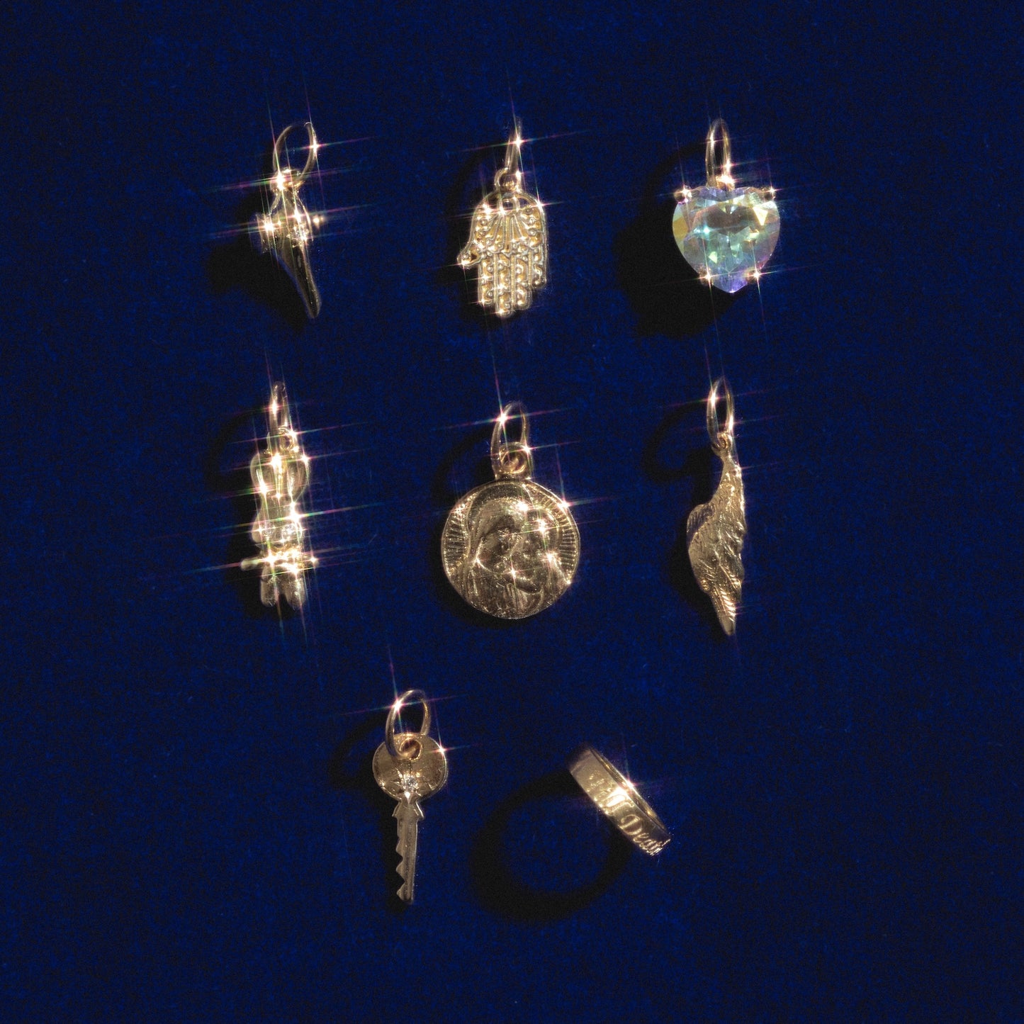 Eight whimsical 14k solid yellow gold charms and a ring from 14k Charms by Charlie and Marcelle, including shapes like a heart, key, angel wings, and hand, arranged on blue velvet with sparkling golden shine.