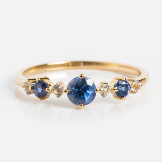 The Solid Gold 2024 December Capsule Classic Tanzanite Ring (Sample Size 5) features a round tanzanite centerpiece with smaller tanzanites and white topaz accents, set against a white background.