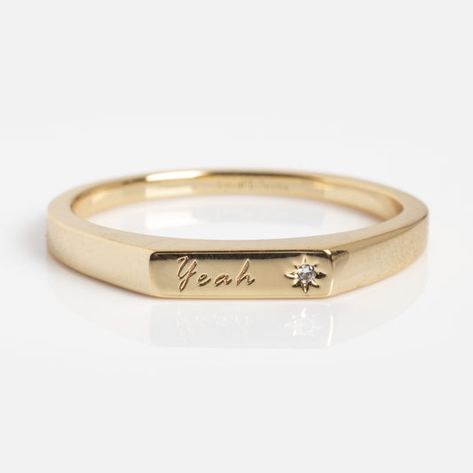 The Tiny Conversation Stacking Ring is a gold band engraved with "Yeah" and accented by a small sparkling gemstone next to the inscription, shown on a white background.
