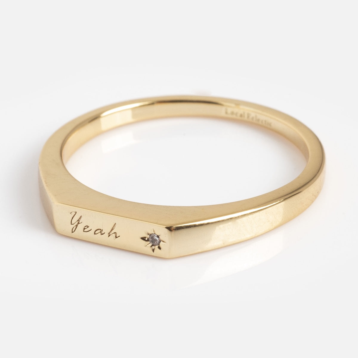 The Tiny Conversation Stacking Ring is a gold ring with a flat face engraved with “Yeah” and a small gemstone beside the engraving, shown on a plain white background.