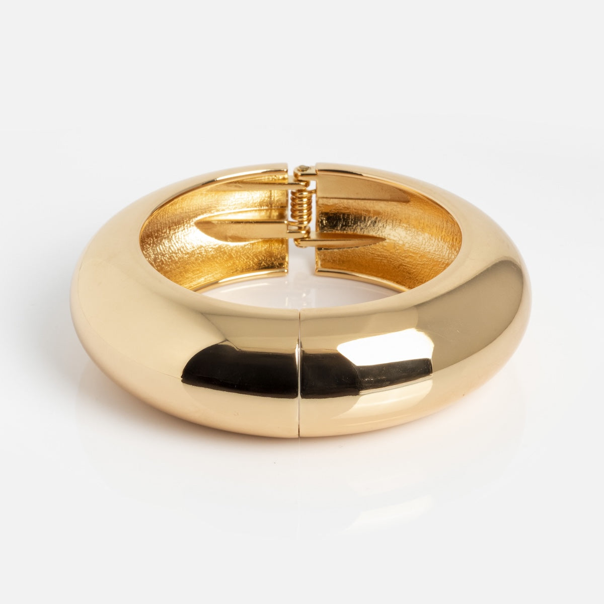 The Bangle Bracelet is a chunky, round gold accessory with a glossy finish and hidden hinge clasp, displayed closed on a white surface.