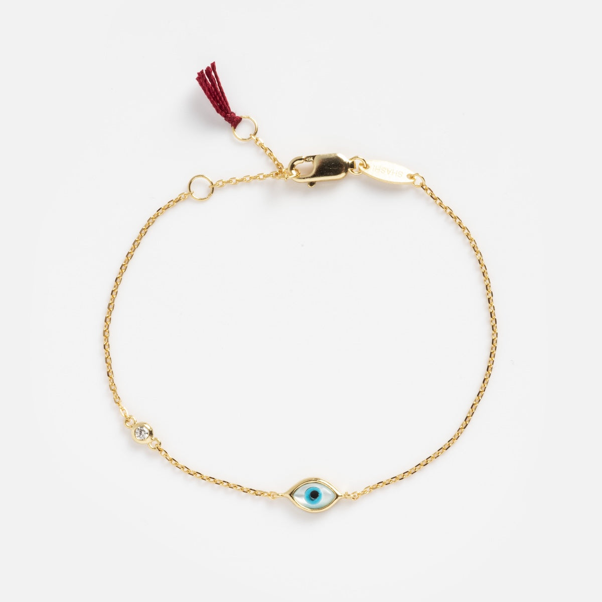 The Madison Evil Eye Bracelet is a delicate 14k vermeil piece with a blue evil eye charm, clear gemstone, lobster clasp, and burgundy tassel—blending stylish appeal with protective symbolism.