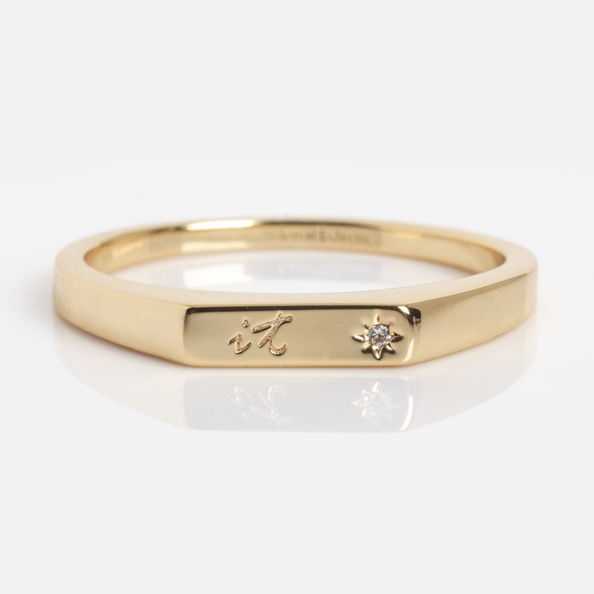 The Tiny Conversation Stacking Ring is a thin gold band engraved with "it" and features a small, star-set diamond on a flat section, shown against a plain white background.