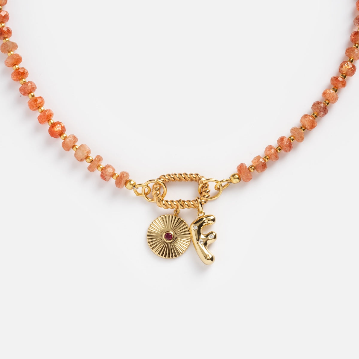 The Customizable Beaded Gemstone Charm Holder Necklace features vibrant orange beads, a gold clasp, a round sunburst charm, and a small gold figure charm.