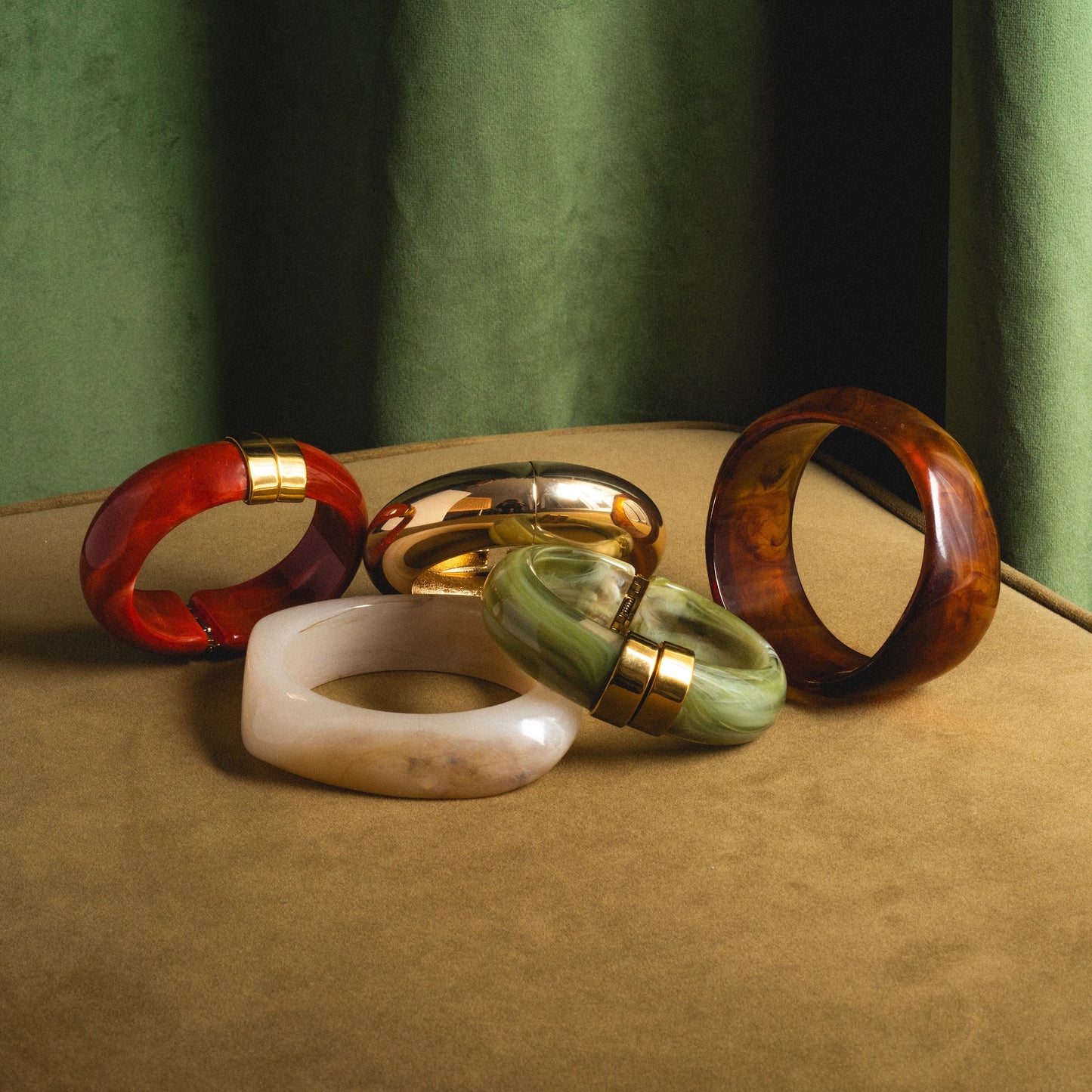 Five Bangle Bracelets in red, gold, green, white, and amber are displayed on a brown surface with a green curtain backdrop. Some Bangle Bracelets feature gold accents.
