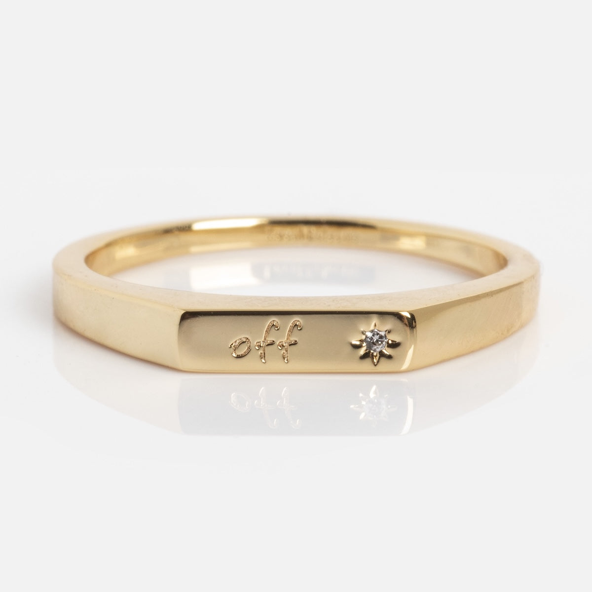 The Tiny Conversation Stacking Ring is a slim gold band featuring a flat section engraved with "off" and a small round diamond beside the engraving, shown against a plain white background.
