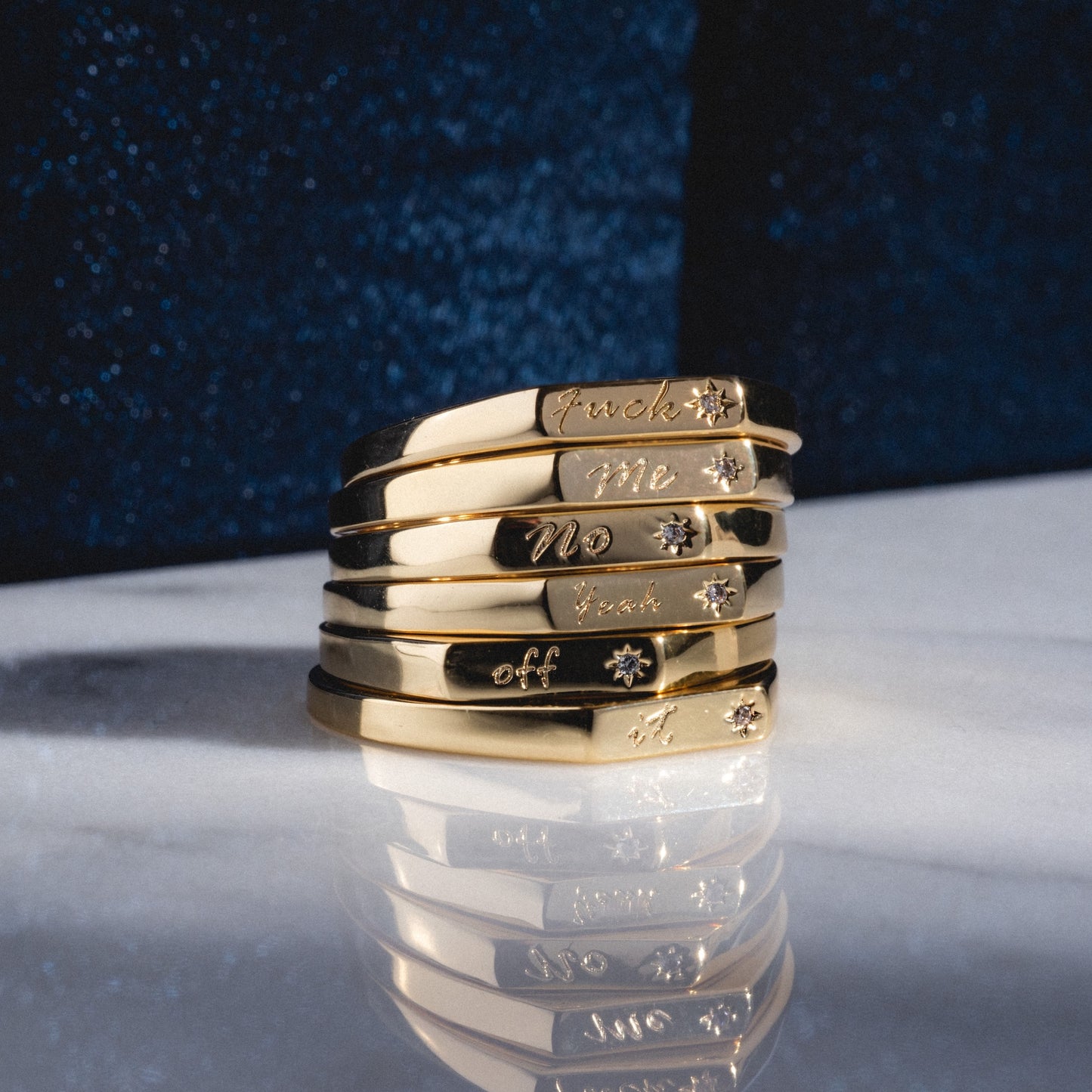 The Tiny Conversation Stacking Ring features six gold bands engraved with “Fuck,” “Me,” “No,” “Thank,” “You,” and “Off,” accented by tiny star or flower motifs, displayed on white marble with a dark blue background.