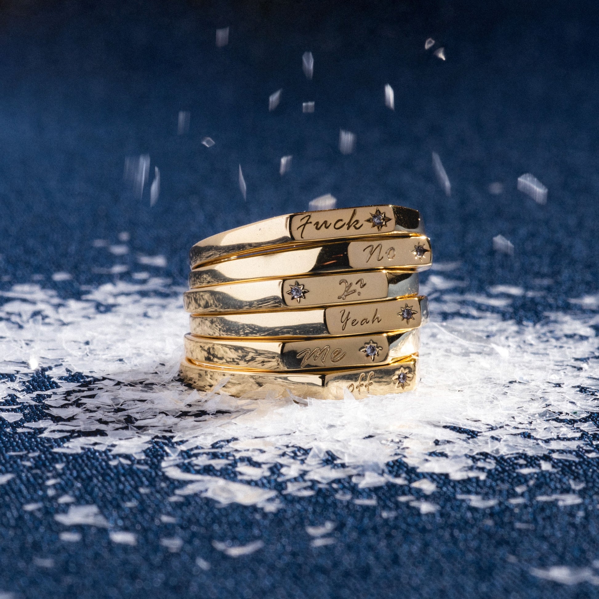 The Tiny Conversation Stacking Ring, engraved with bold phrases like “Fuck,” “No,” “Yeah,” and “Me,” is displayed in a stack of five atop denim, surrounded by white flakes that resemble snow.
