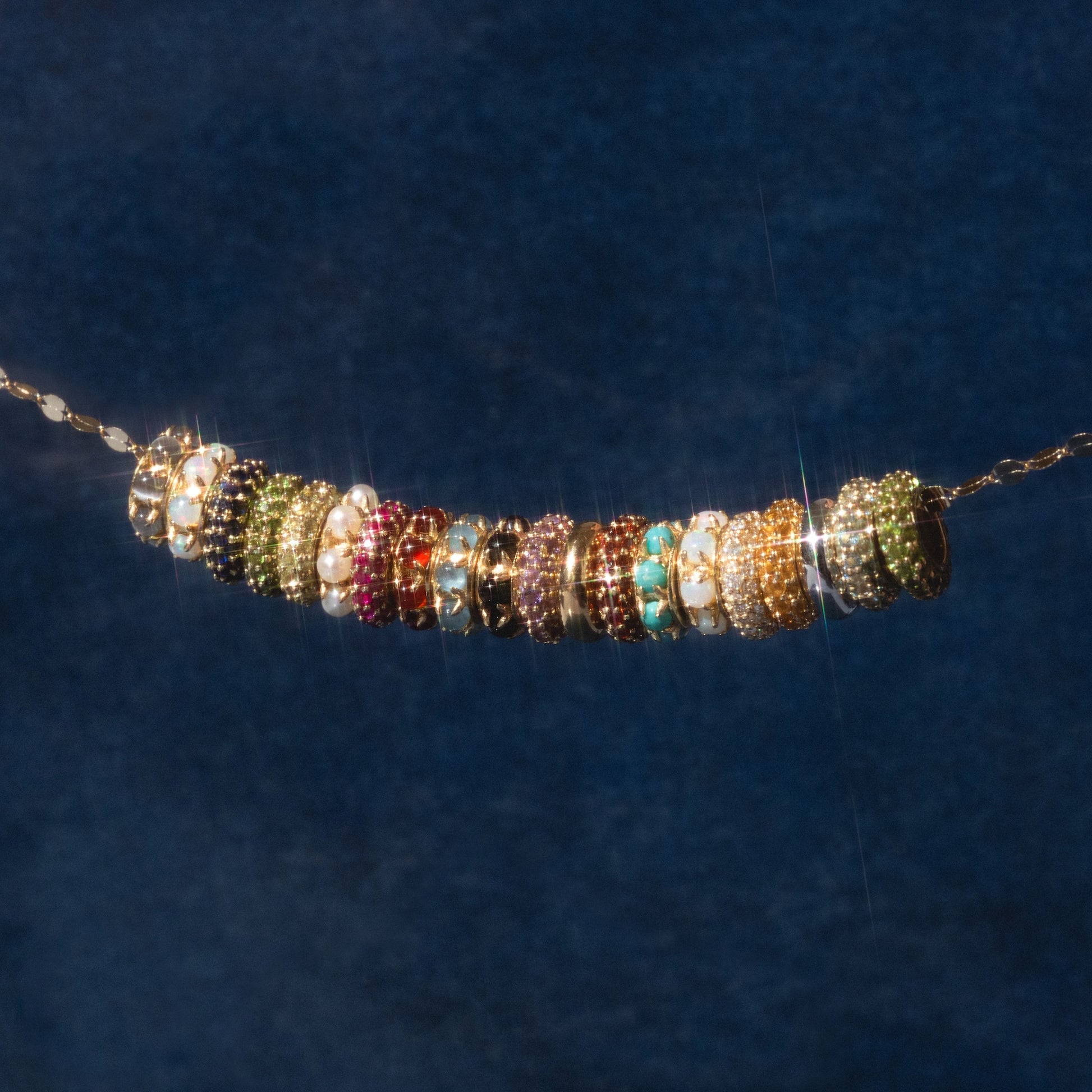 The Solid Gold Anniversaire Charm is a gold chain holding colorful beaded rings with unique designs and gemstones—featuring pearls, turquoise, and sparkling stones in green, red, orange, and black—set against a dark blue background.