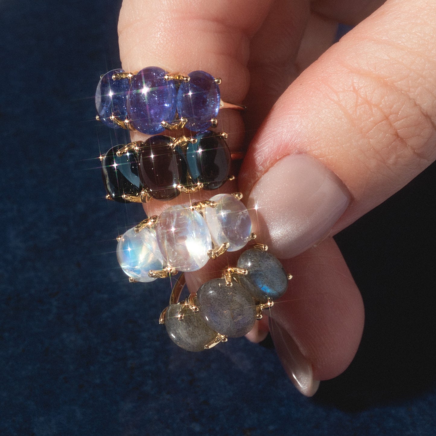 A hand with light nails holds four Sweetest Gumdrop Rings, each in 14k solid yellow gold and featuring three large, oval natural gemstones in assorted colors, sparkling against a dark blue background.