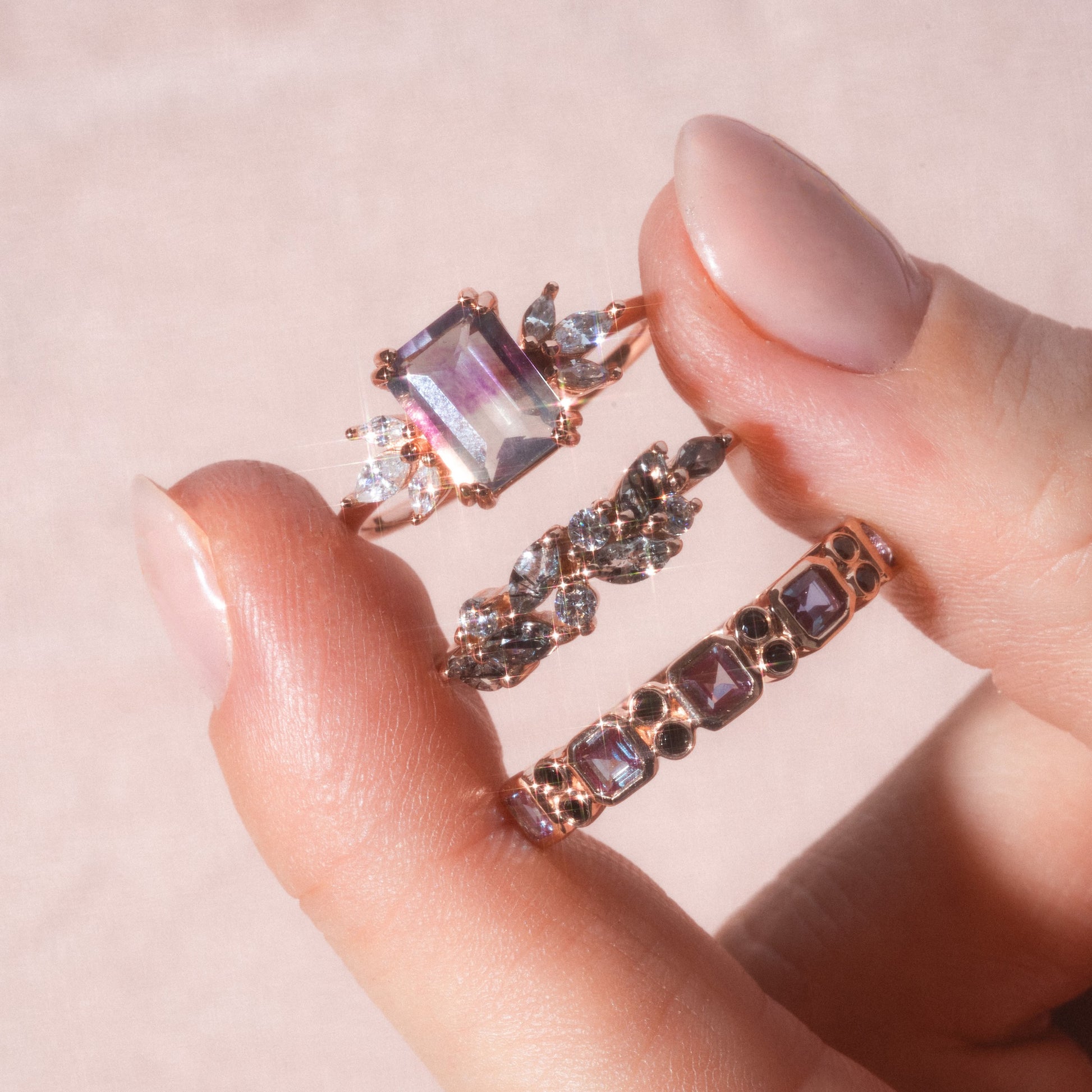 A hand with light pink nails holds three rose gold rings, featuring the 9kt Rose Gold Alexandrite & Black Diamond Juniper Ring alongside others with unique gemstone shapes—rectangular, marquise-cut, and square-cut stones.