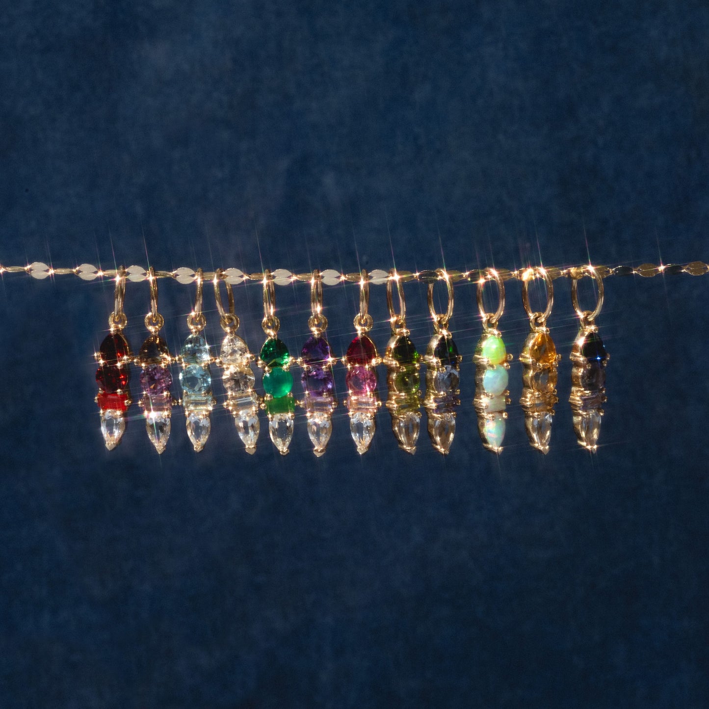 Twelve Solid Gold Modern Ombre Birthstone Charms with gold loops hang on a thin wire against a dark blue background, arranged in a sparkling, colorful spectrum.